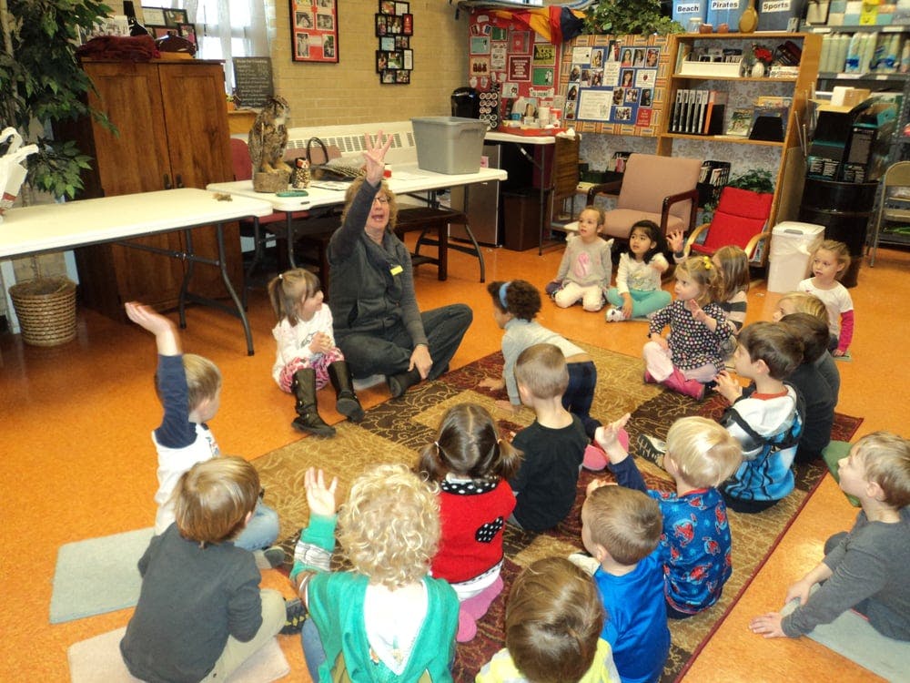Adult and children sitting on a rug with hands raised in a classroom