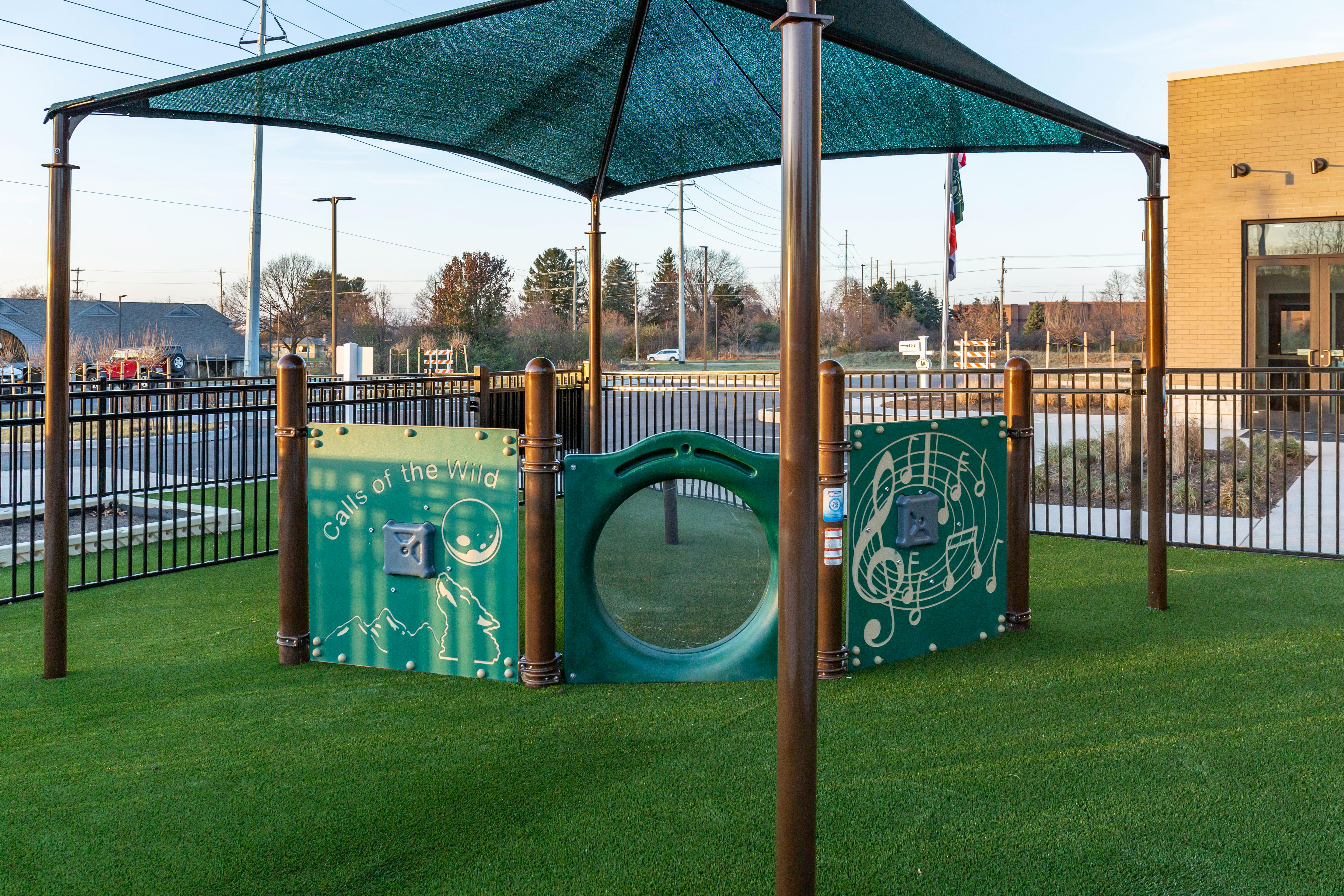 Playground equipment with a green shade and artificial grass, surrounded by a fence