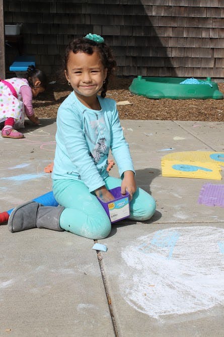 Child kneeling on pavement with chalk, holding a container