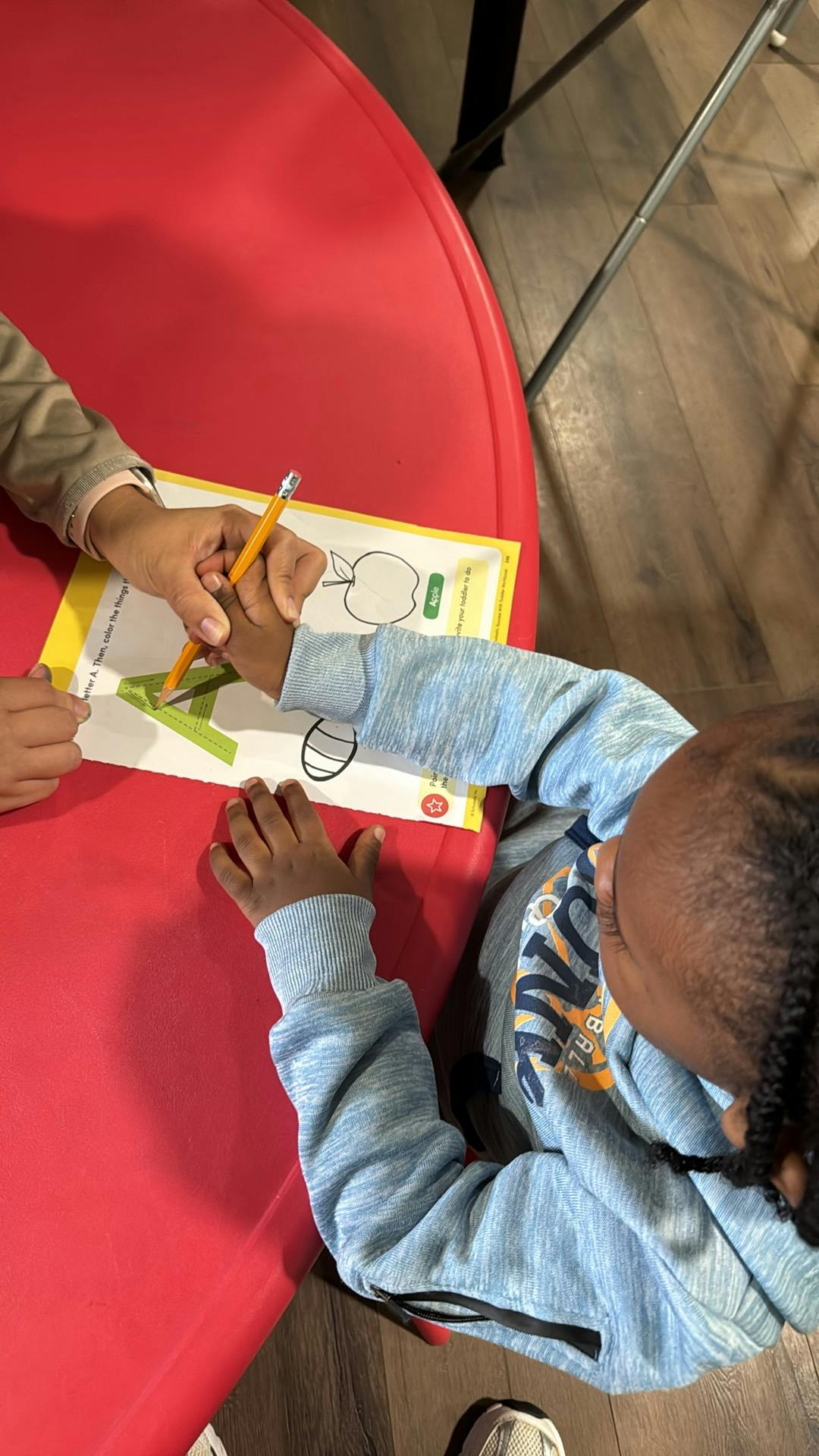 Adult helping a child hold a pencil to color the letter A on a worksheet