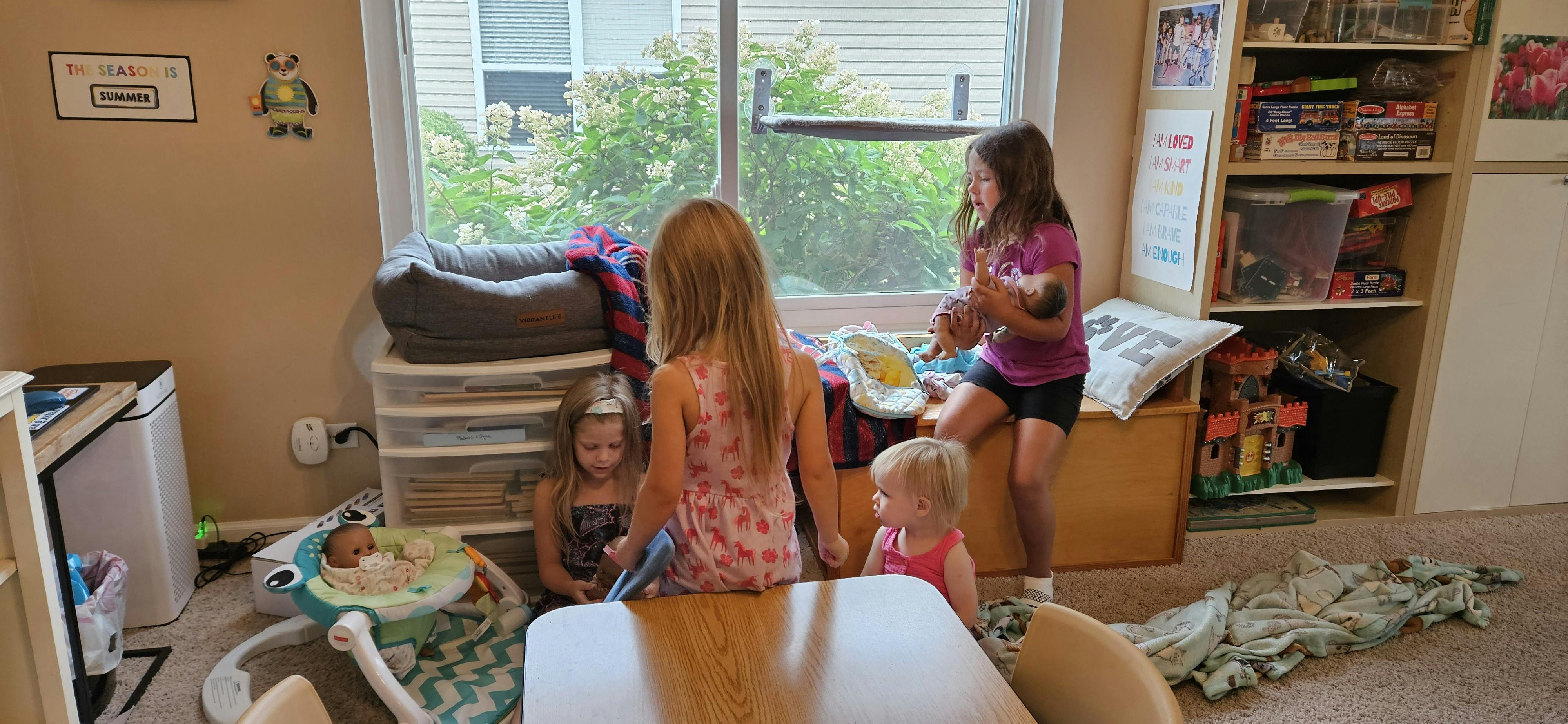 Four children in a room, one holding a doll, another in a baby bouncer