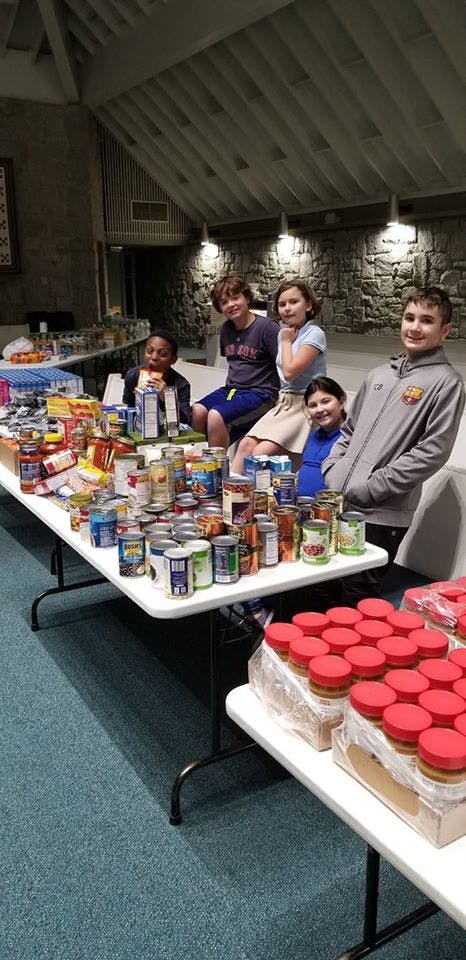 Five children sitting behind tables filled with canned goods and jars
