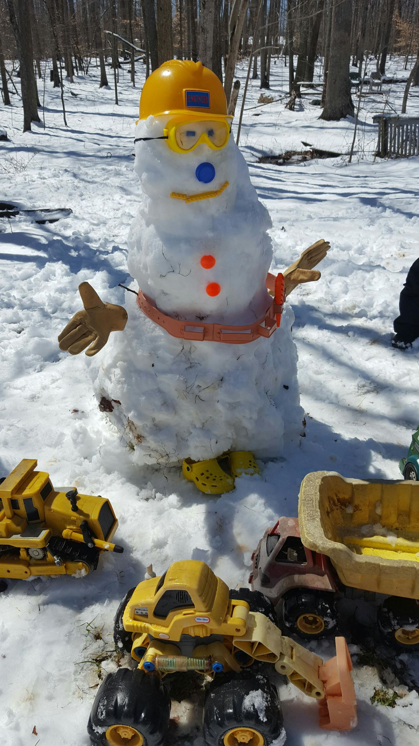 Snowman wearing a yellow hard hat and goggles with toy trucks in the snow
