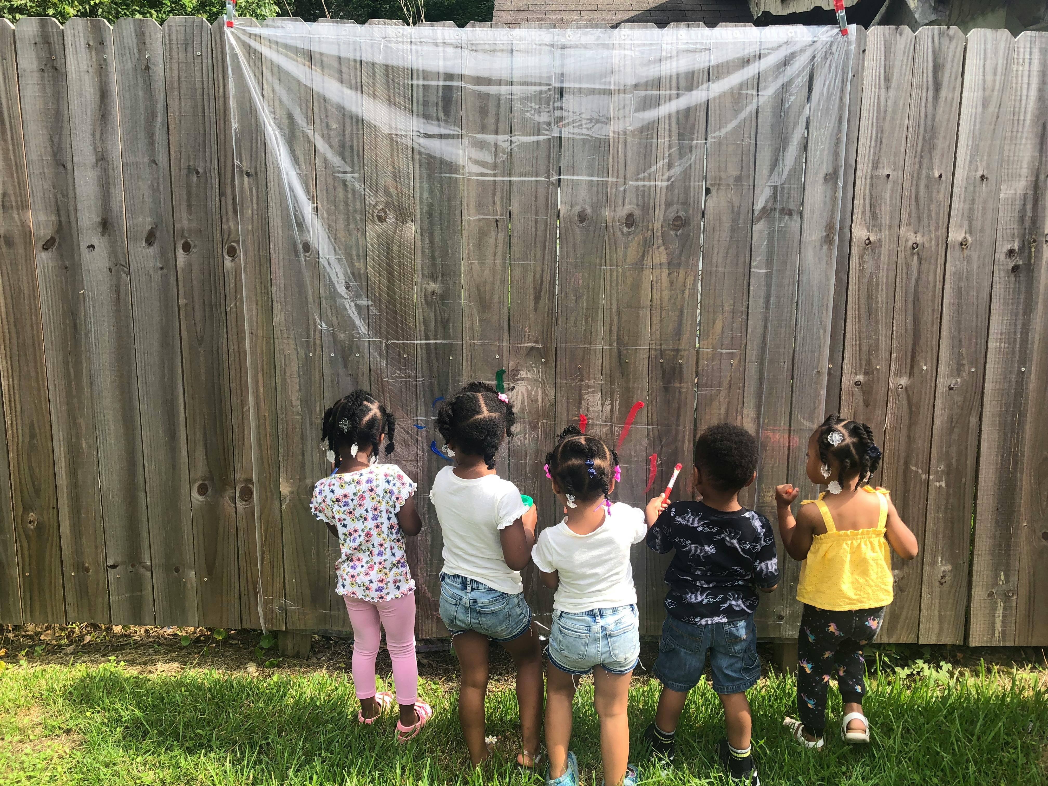 Five children painting on a clear sheet attached to a wooden fence