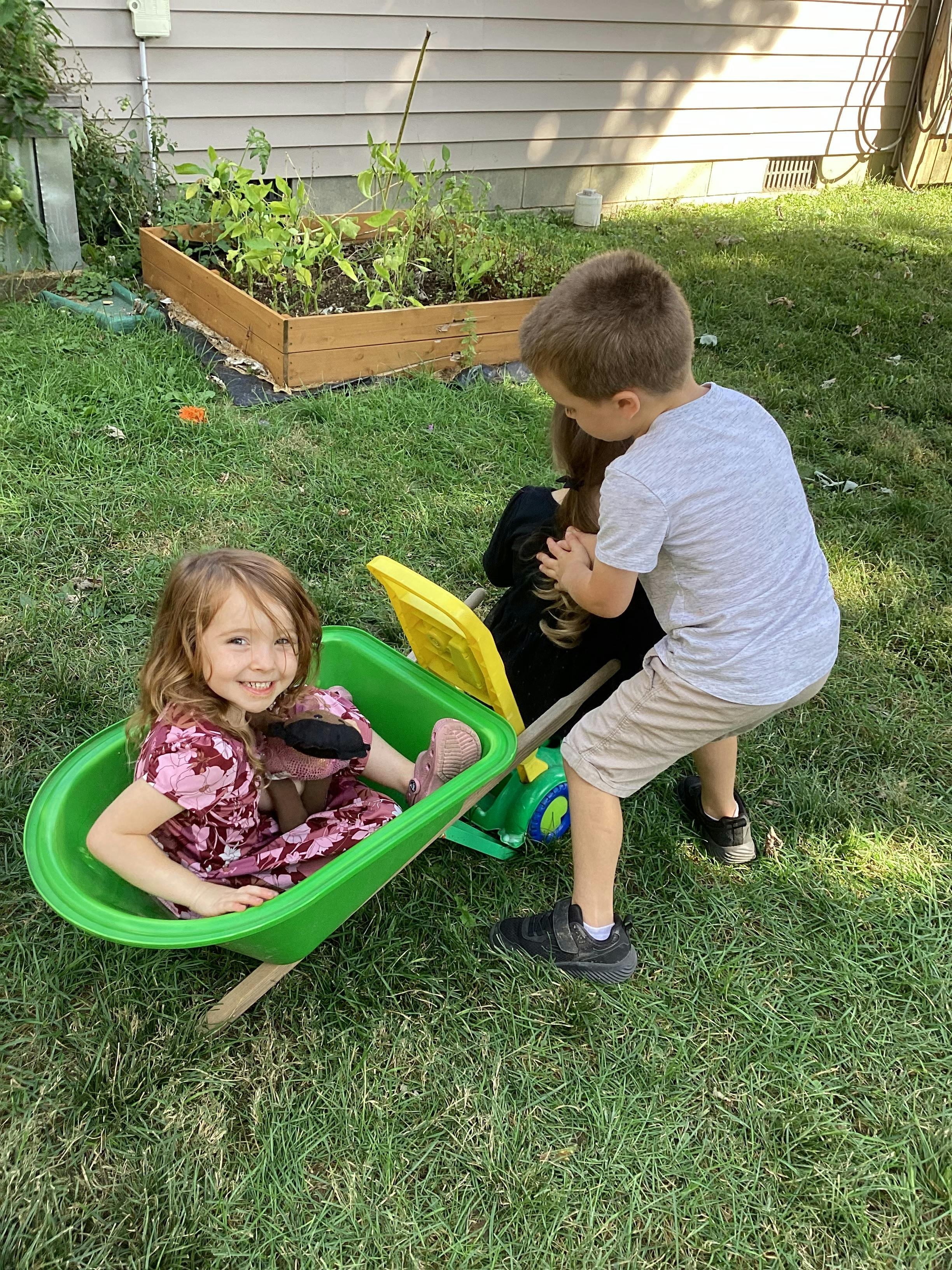 Child sitting in a green toy wheelbarrow while another child stands nearby
