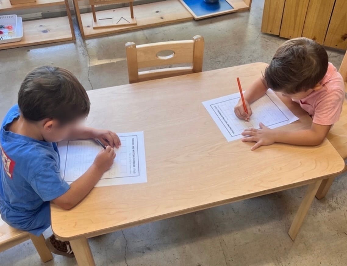 Two children writing on paper at a table in a classroom