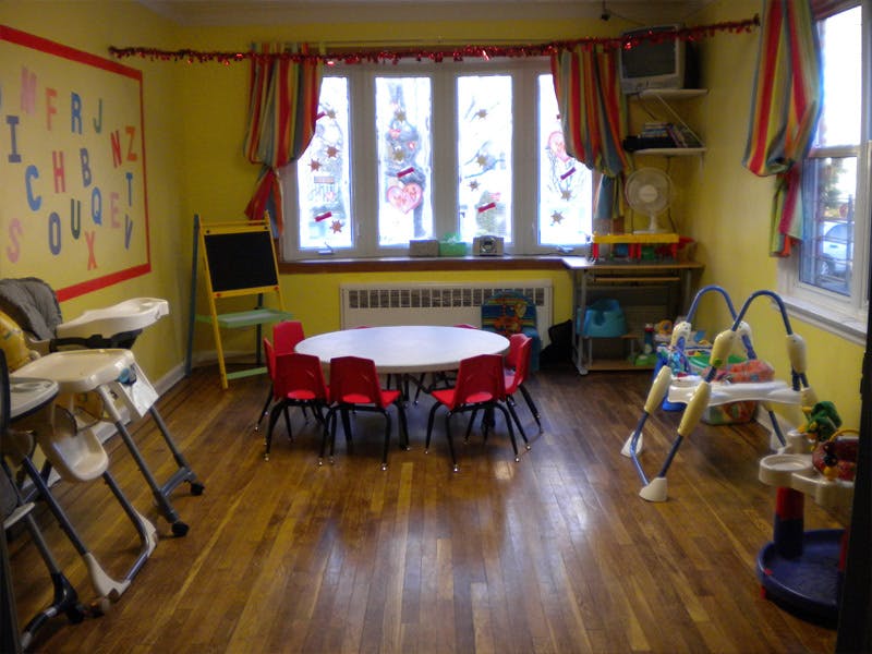 A classroom with a small round table and red chairs, high chairs, and a play mat