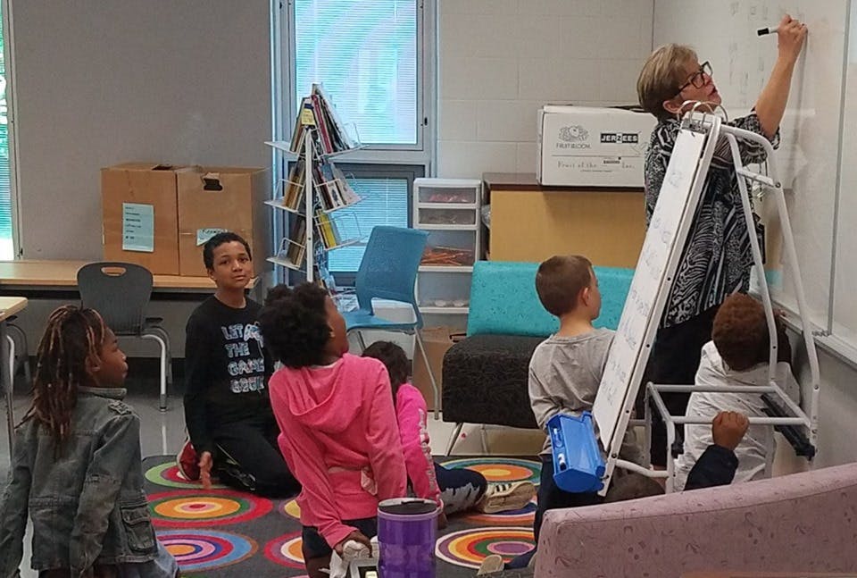 Adult on a ladder writing on a whiteboard with children watching
