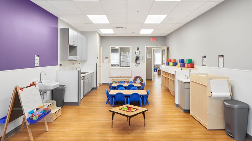 Classroom with small blue tables and chairs, easel, and sink area