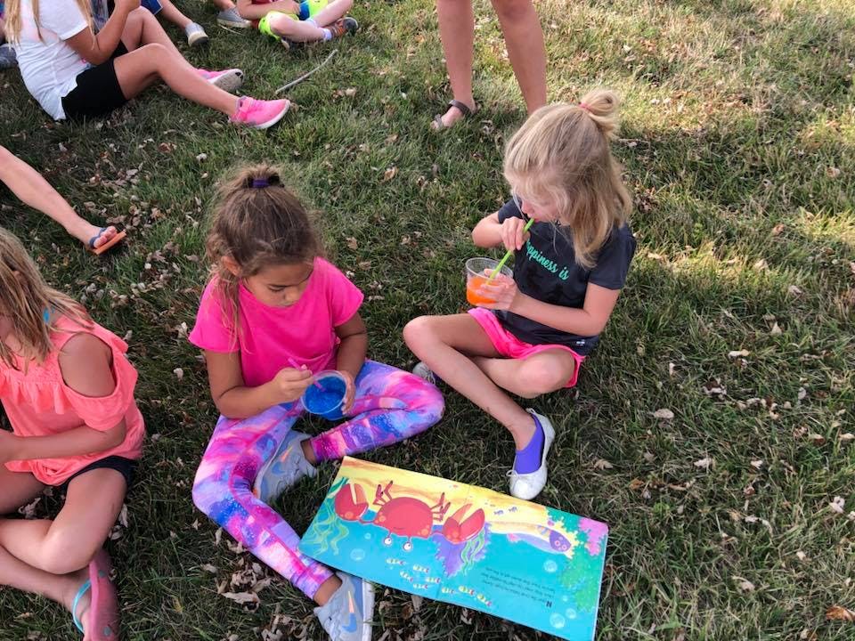 Children sitting on grass, one painting and another drinking from a cup