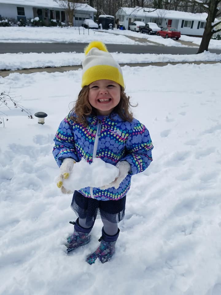 Child in winter clothing holding a snowball in a snowy yard