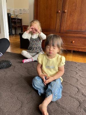 Two children sitting on a rug, one with eyes closed and the other with hands to eyes