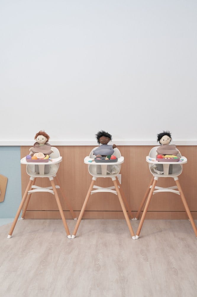 Three dolls sitting in high chairs with play food on their trays