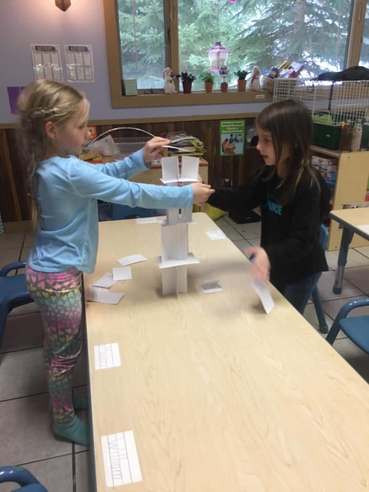 Two children building a tower with paper cups and cards on a table