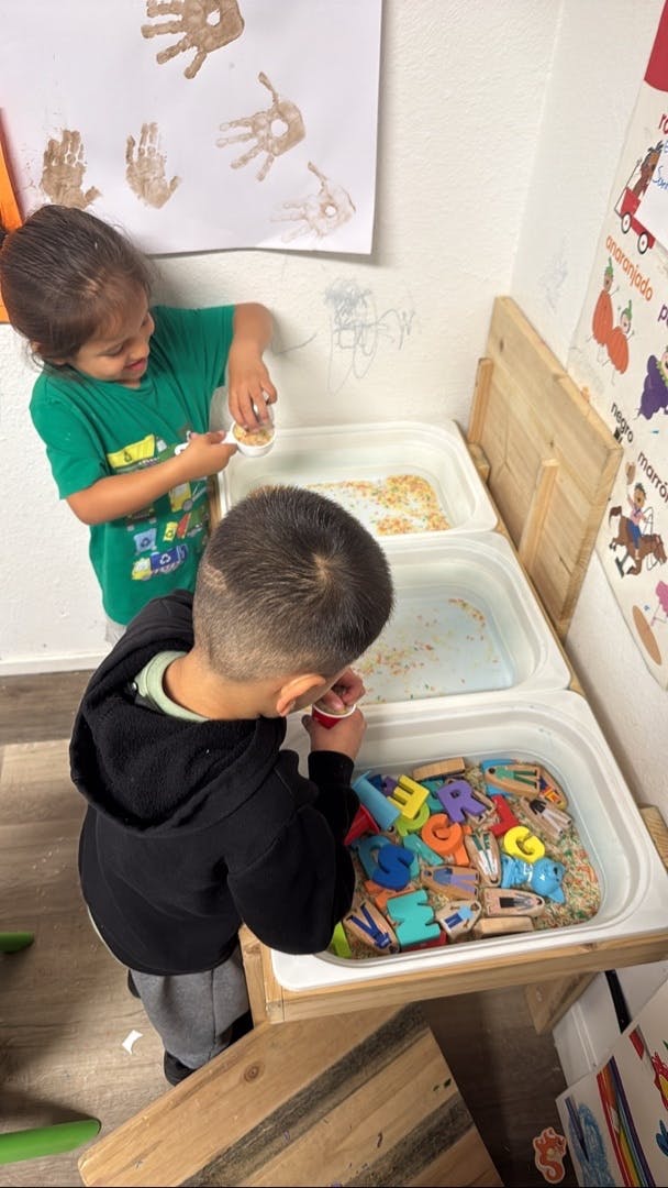 Two children playing at a sensory table with colorful letters and grains