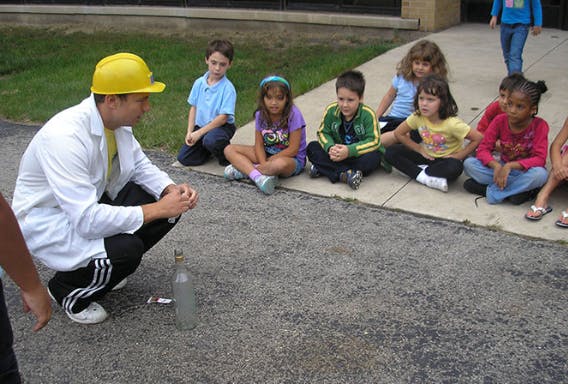Person in lab coat and hard hat crouching in front of children