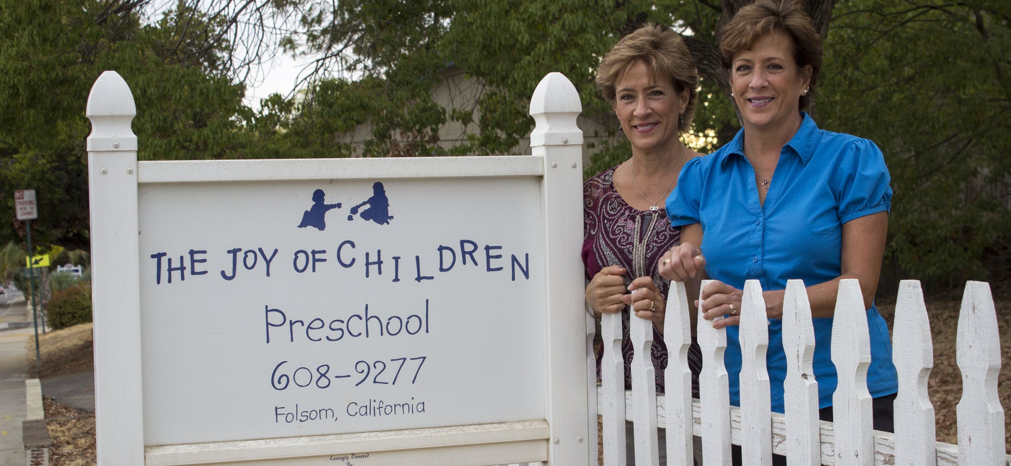 Two women standing behind a white picket fence next to a preschool sign