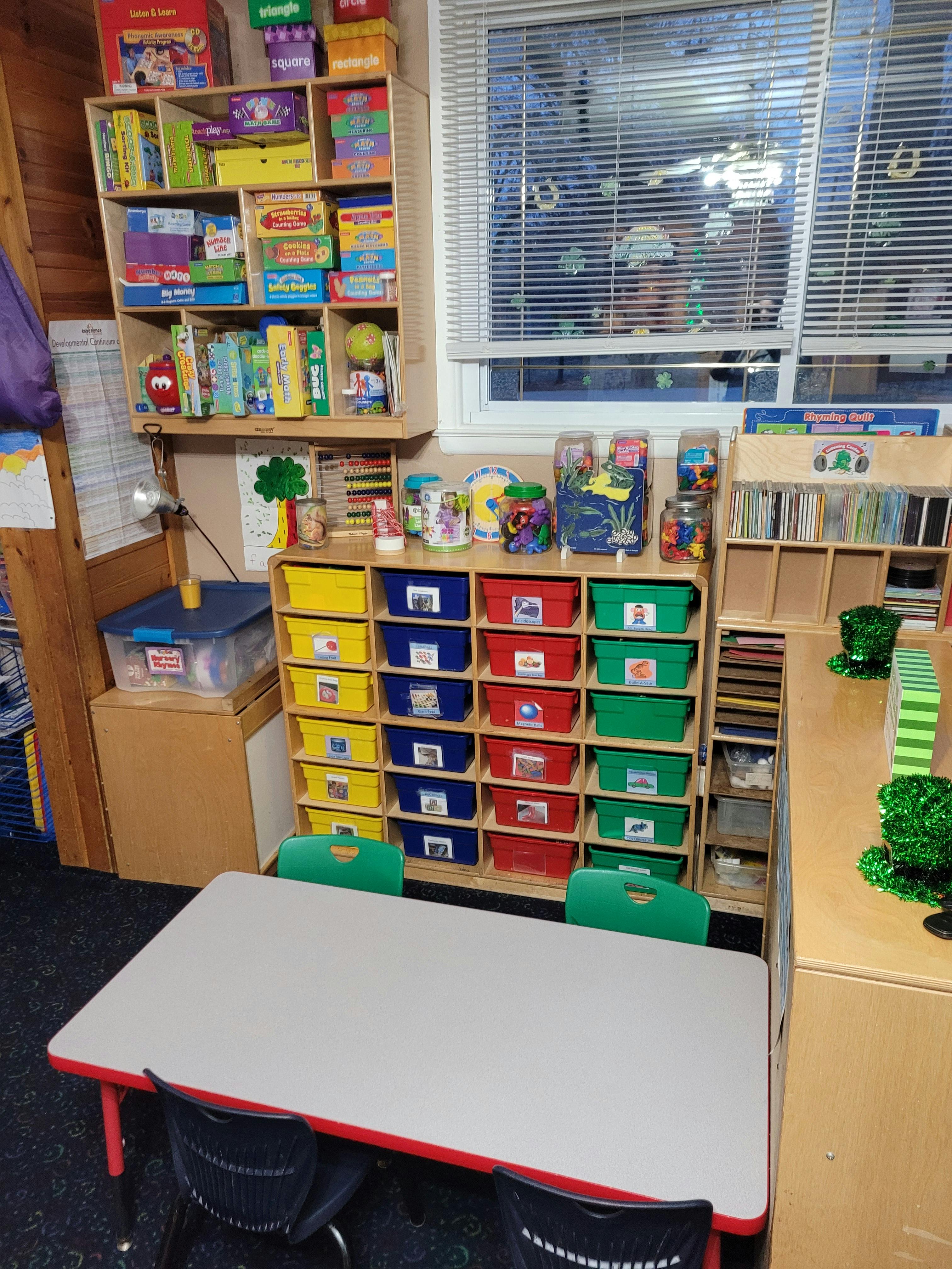 Classroom with shelves full of toys and games, colorful bins, and a small table with chairs