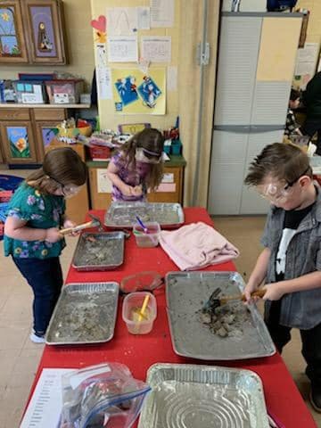 Three children wearing safety glasses working with trays of dirt and tools