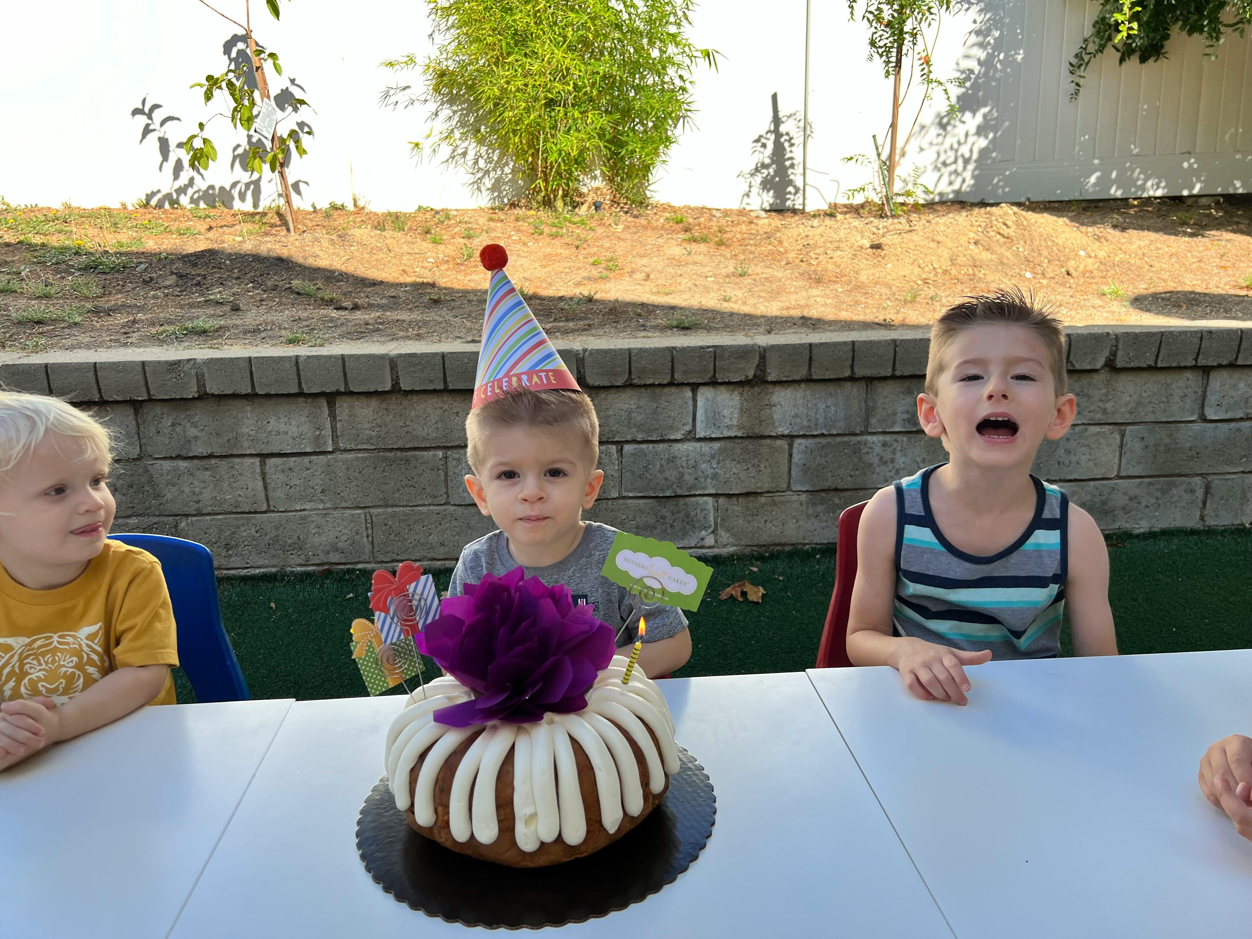 Three children at a table with a cake, one child wearing a party hat