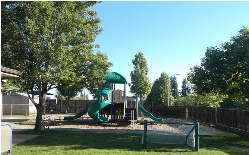A green and tan playground set with slides in a fenced area with trees