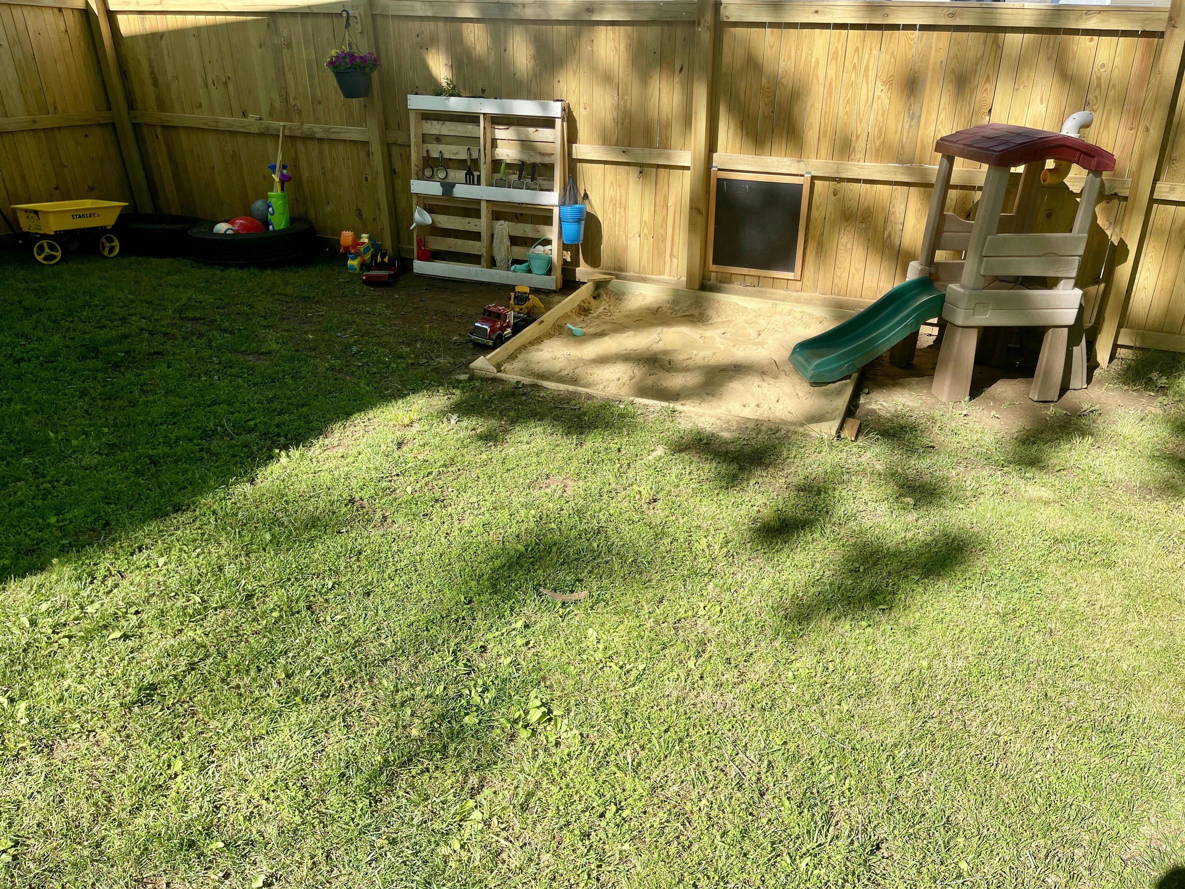 Backyard with a wooden fence, green grass, sandbox, and play structure with slide