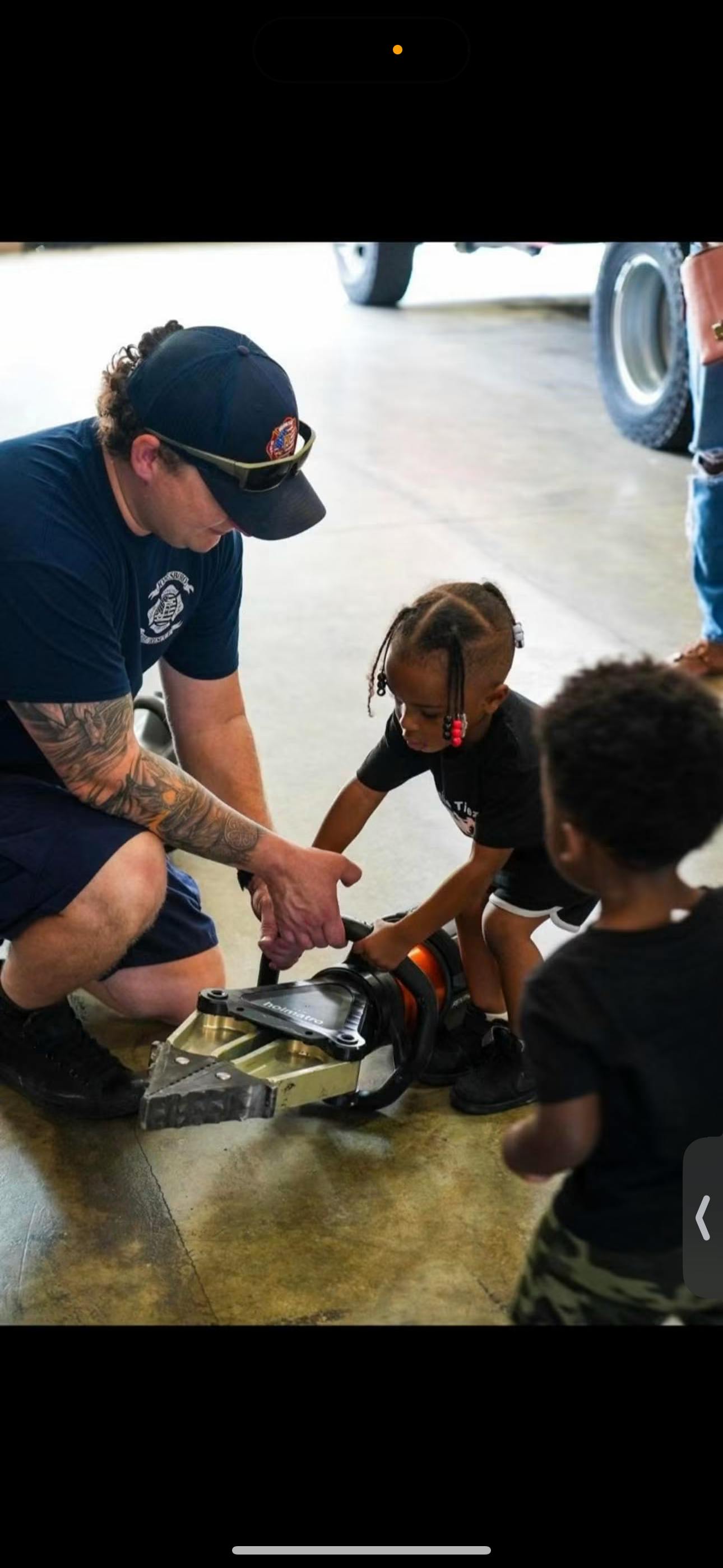 A person helping a child hold a hydraulic rescue tool in a garage