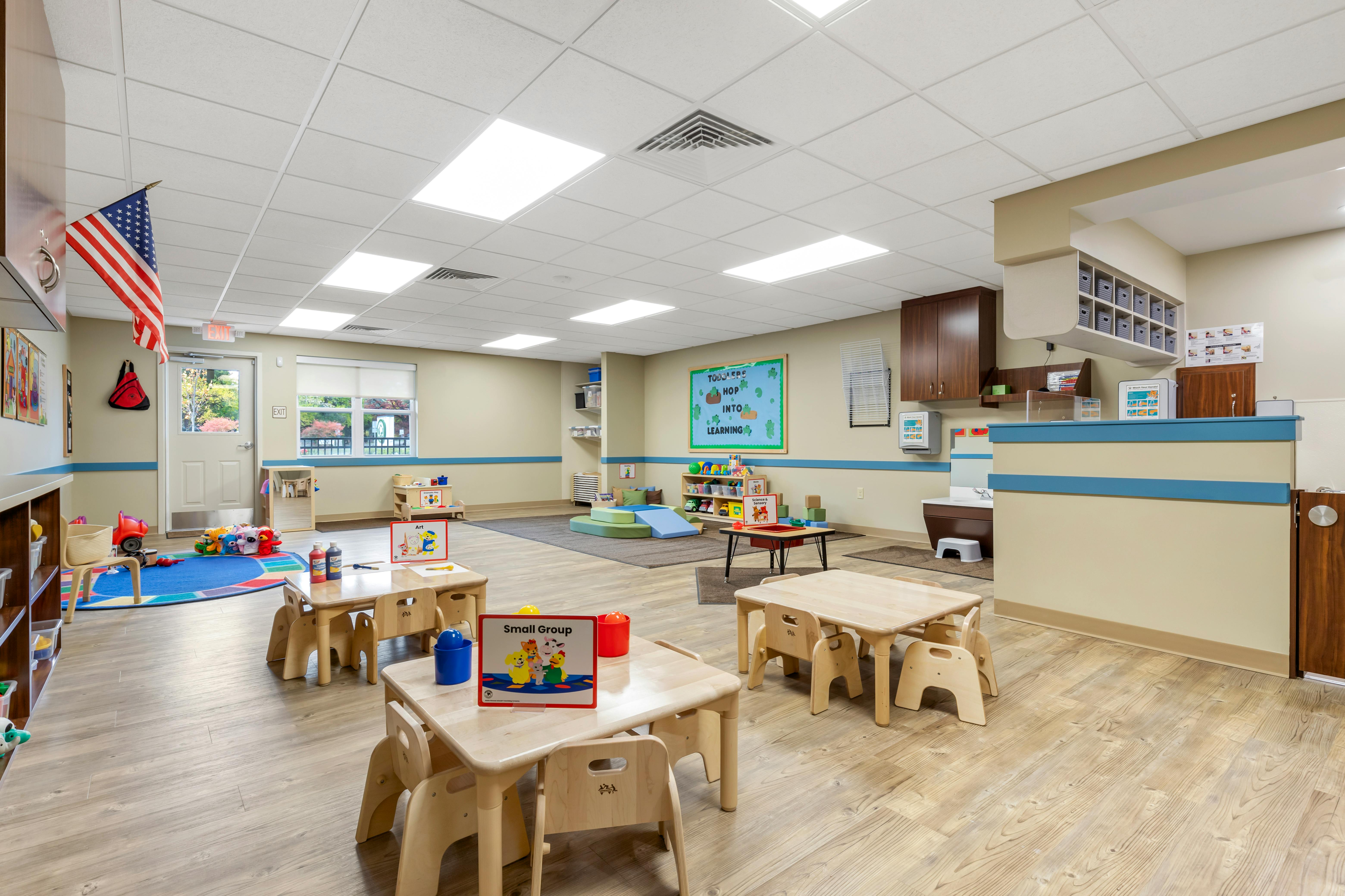 A classroom with small tables and chairs, a play area, and an American flag