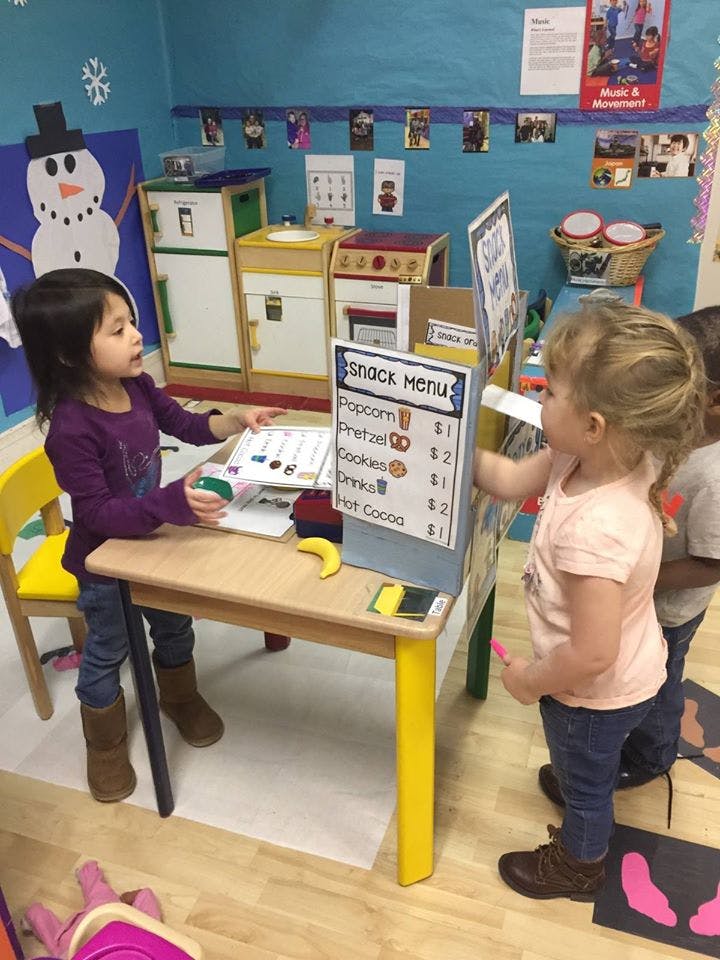 Two children playing with a snack menu at a small table in a classroom