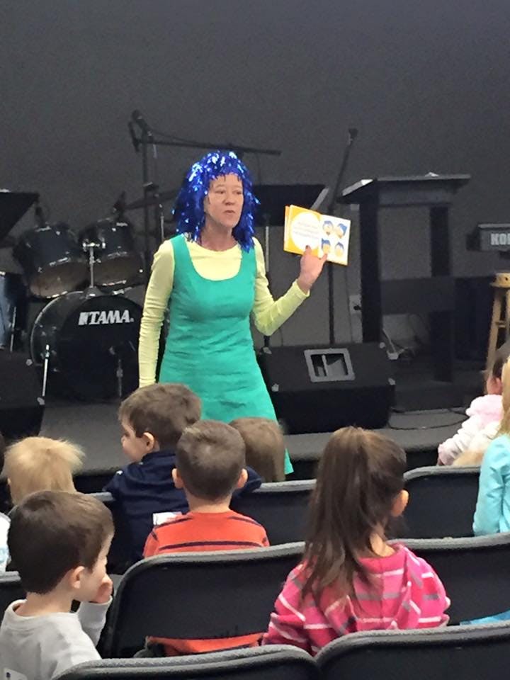 Person in blue wig and green dress holding a book for children