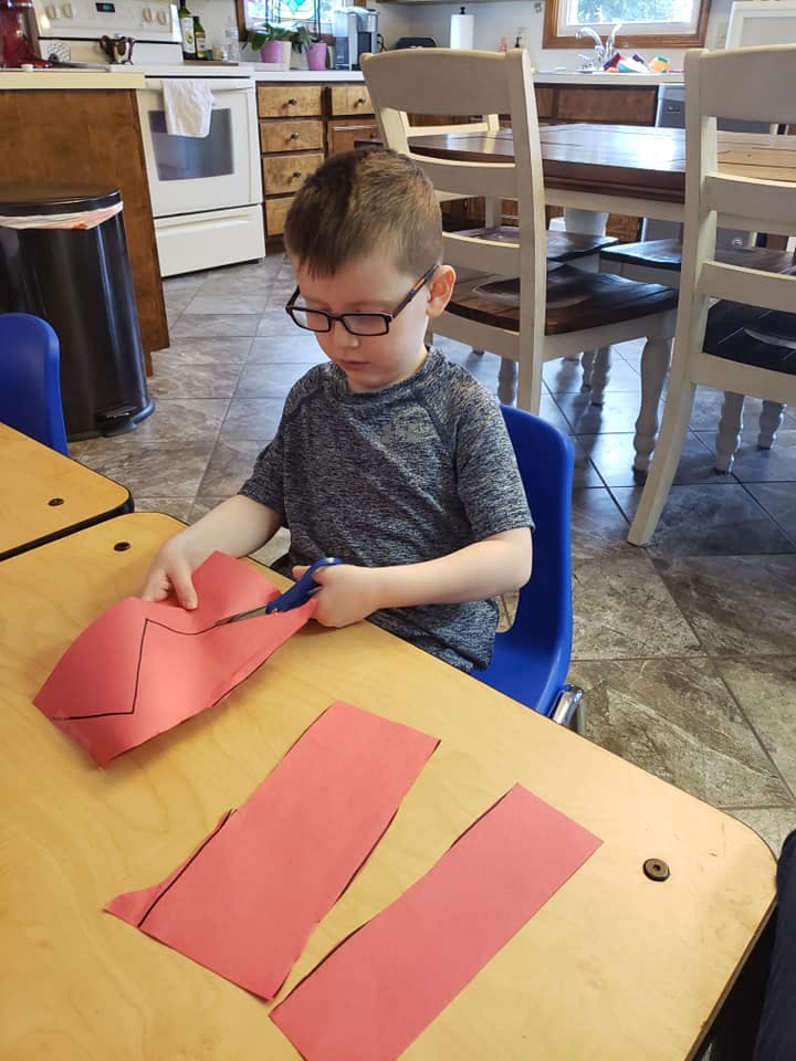 Child in glasses cutting red paper with scissors at a table
