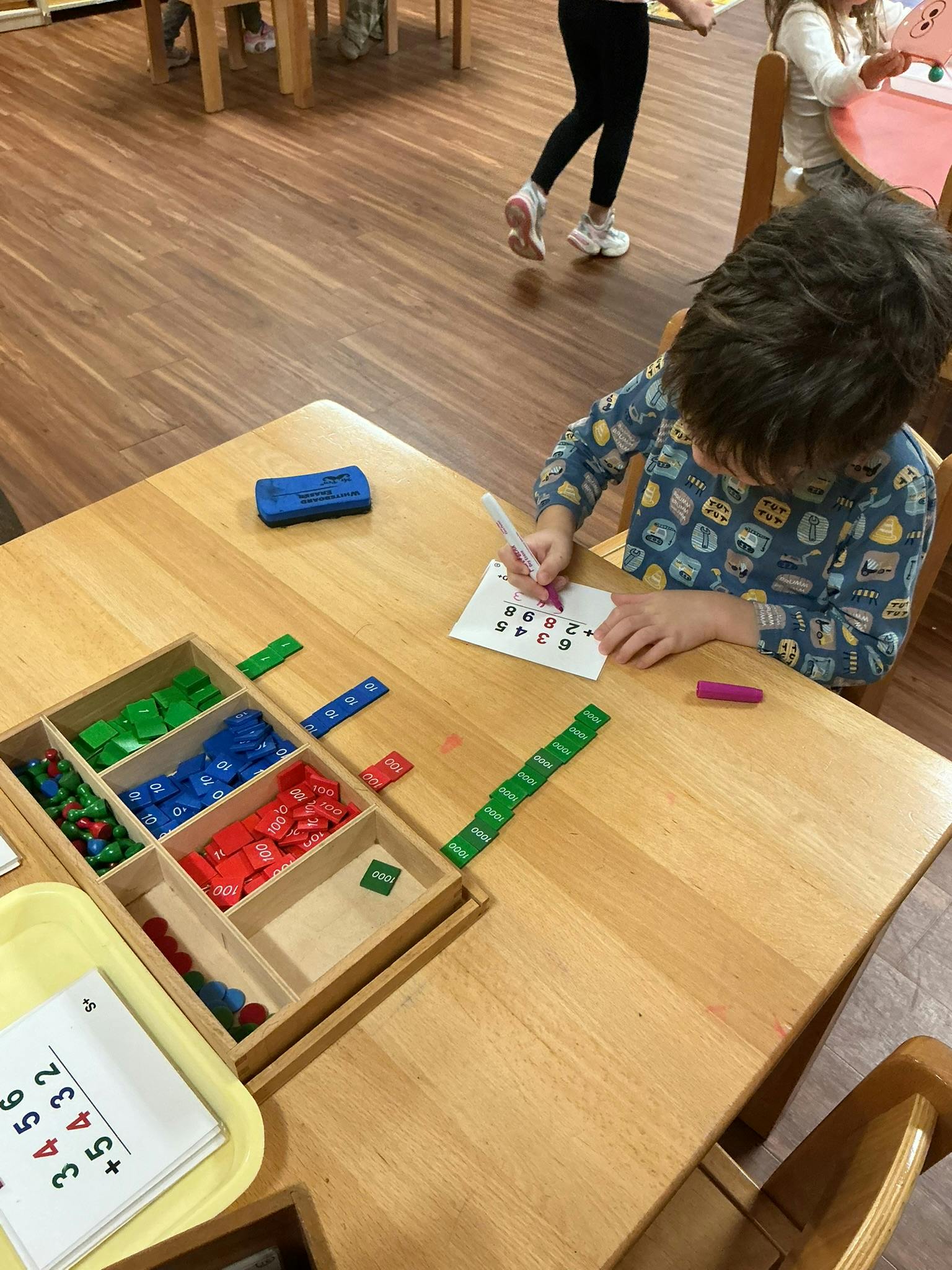 Child writing on paper at a table with math manipulatives