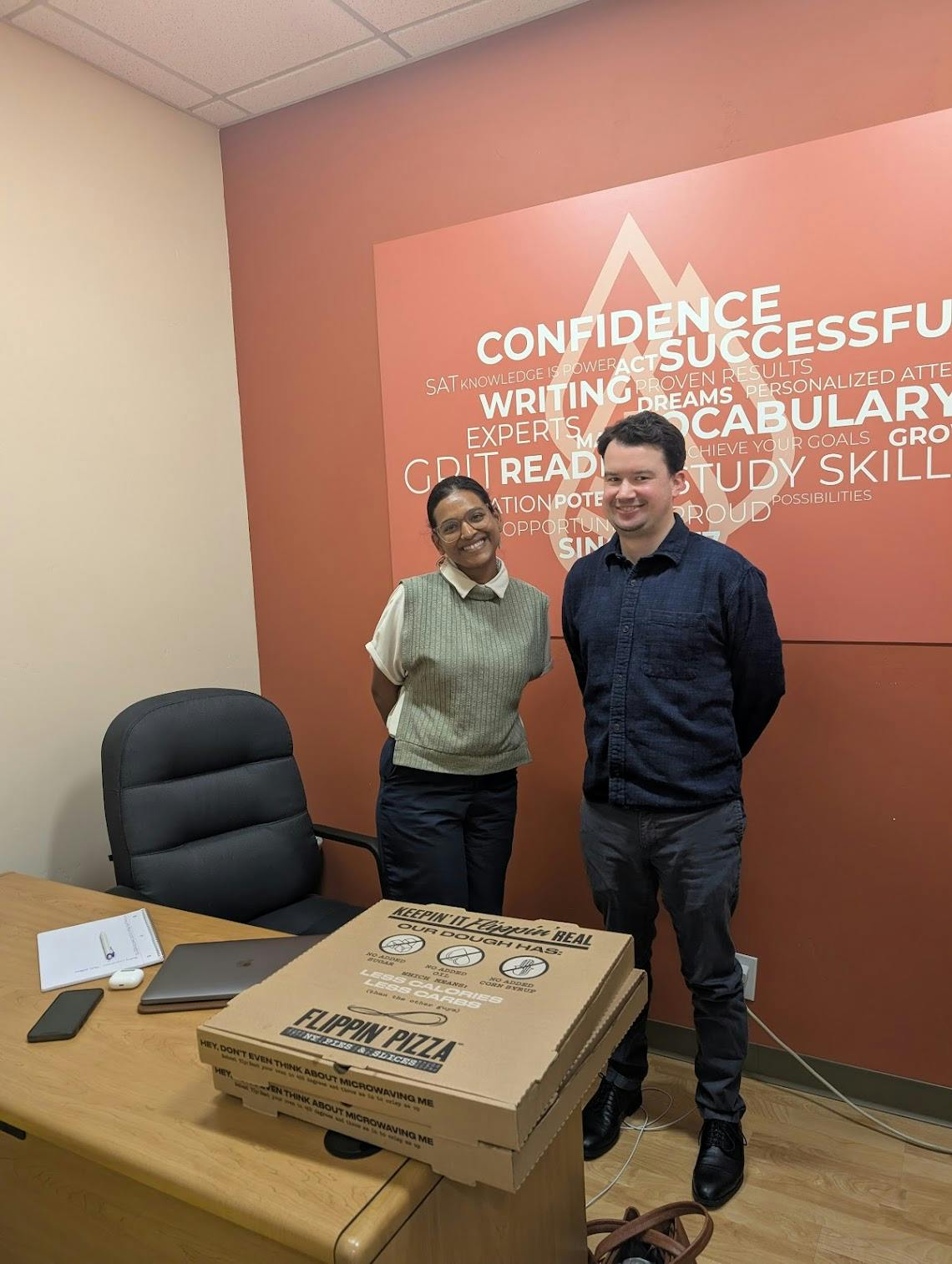 Two people standing next to a stack of pizza boxes on a desk