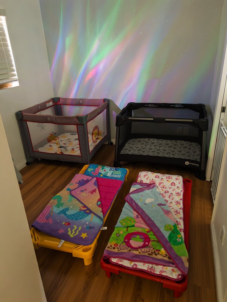 Two playpens and two toddler cots with blankets on a wooden floor