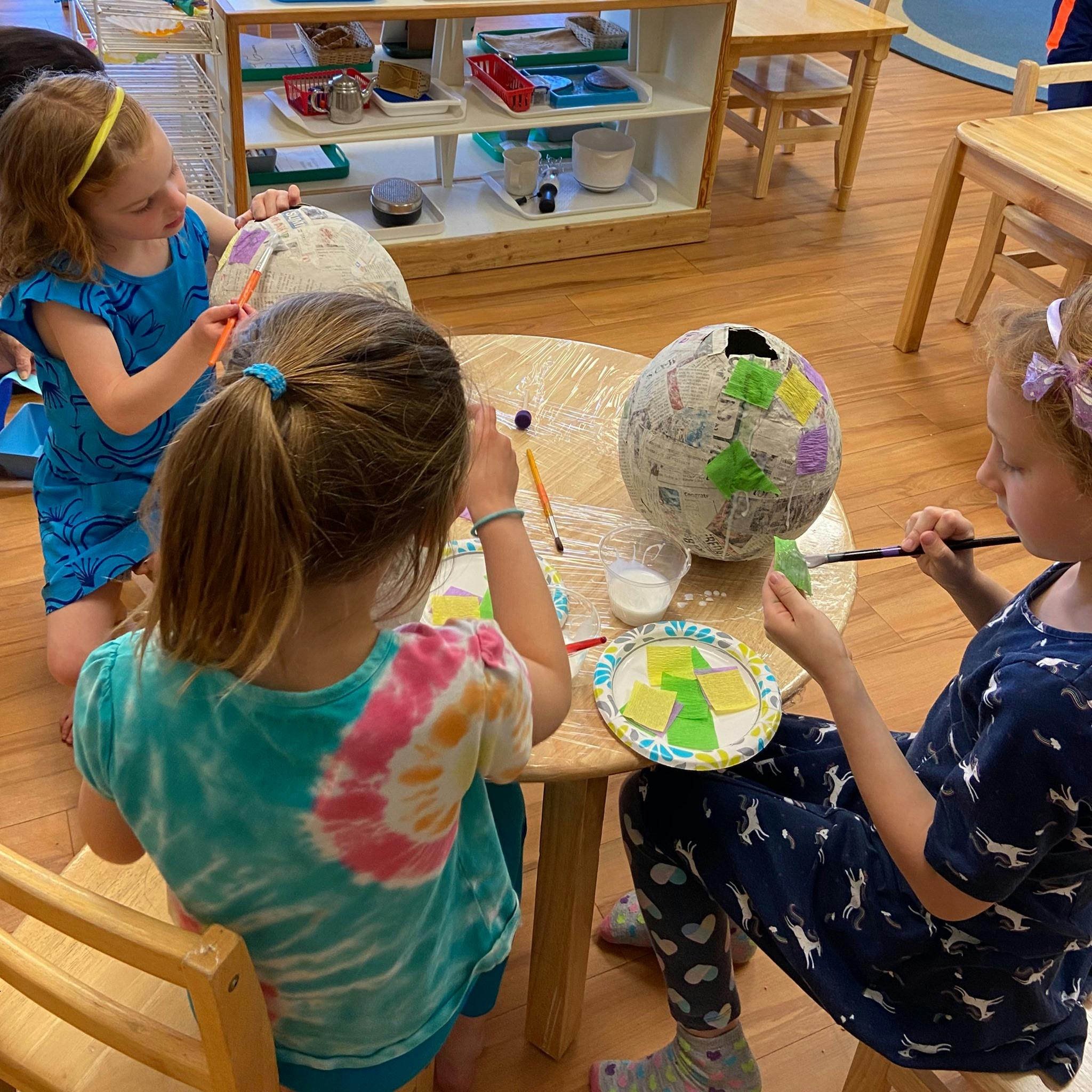 Three children painting paper mache globes at a table