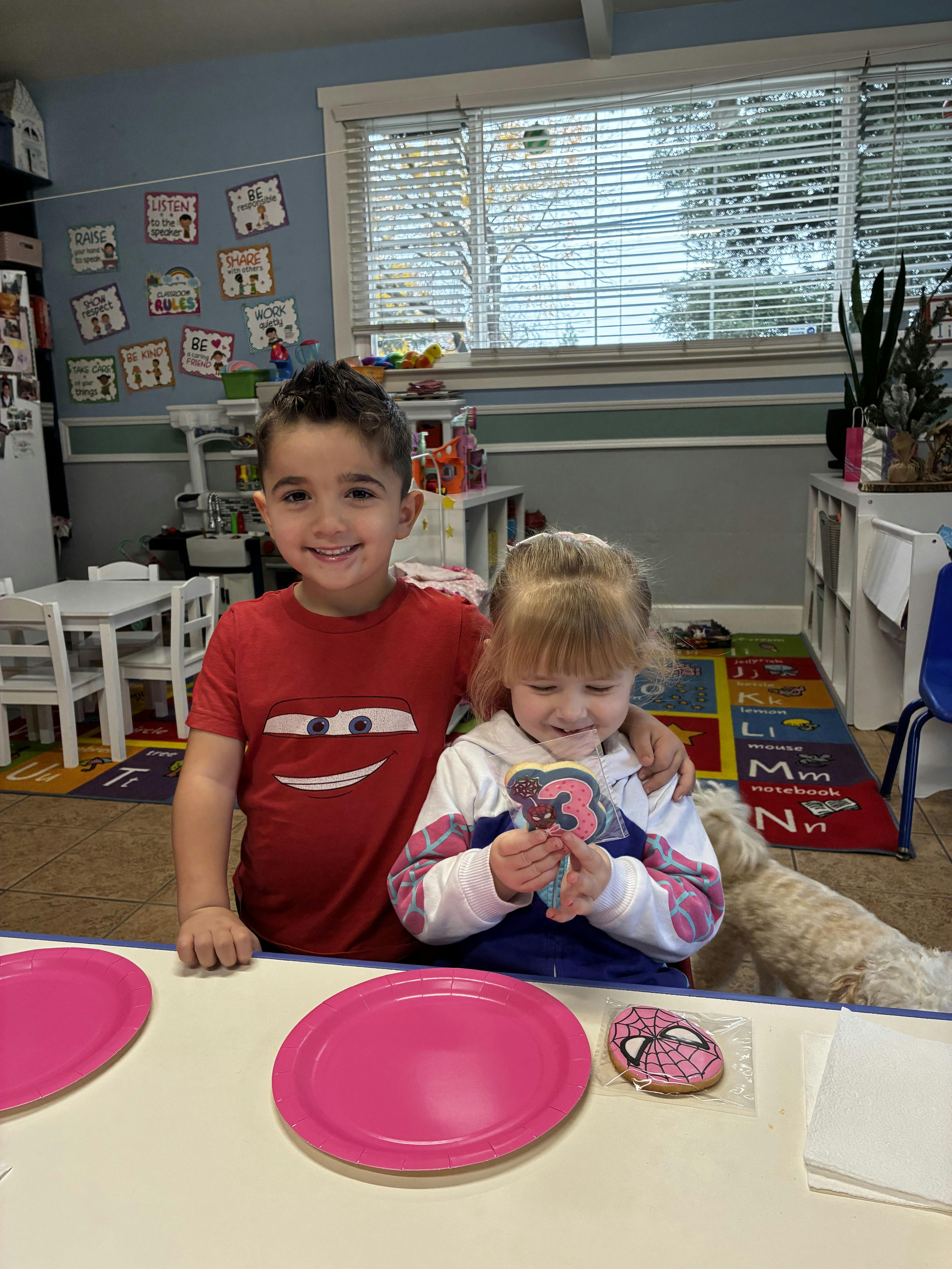 Smiling child with arm around smaller child holding a cookie with a number 3 and spider on it