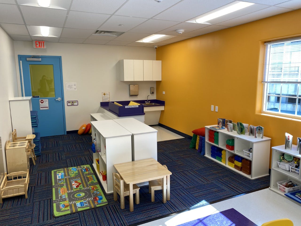 Empty classroom with blue carpet, yellow wall, and small tables and shelves