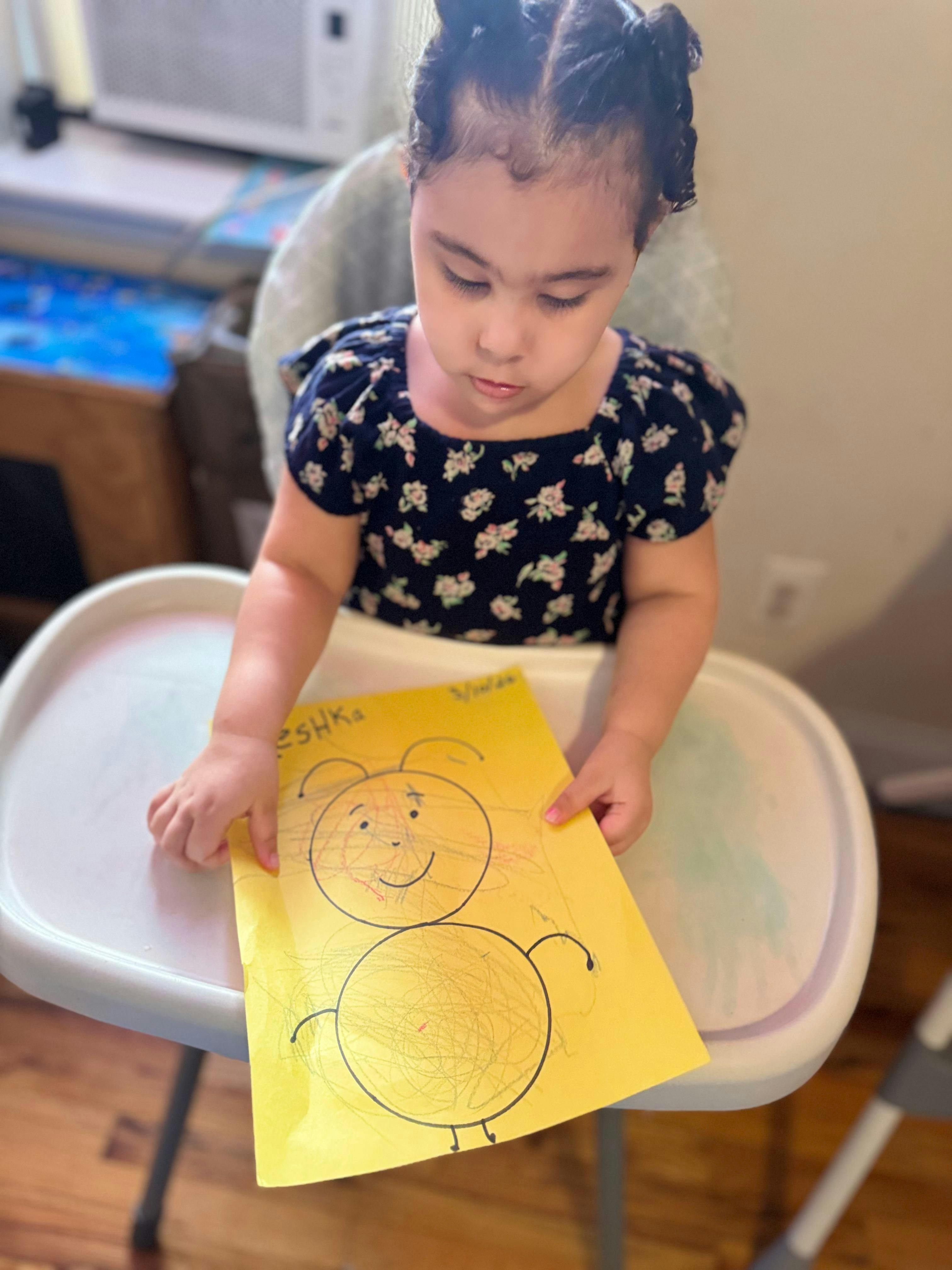 Child in a high chair holding up a yellow paper with a drawing