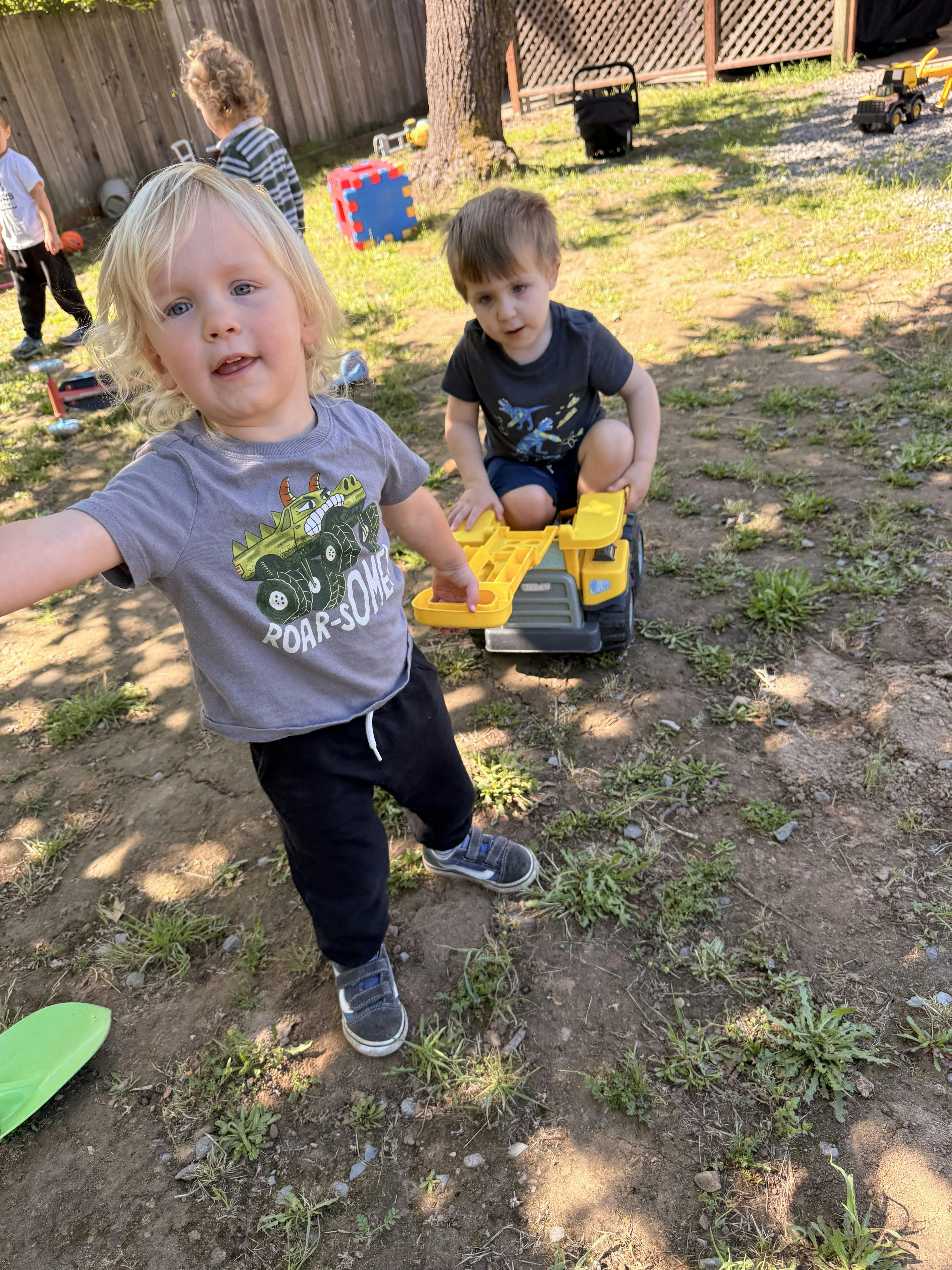 Child in gray shirt holding a toy truck, another child in a black shirt playing