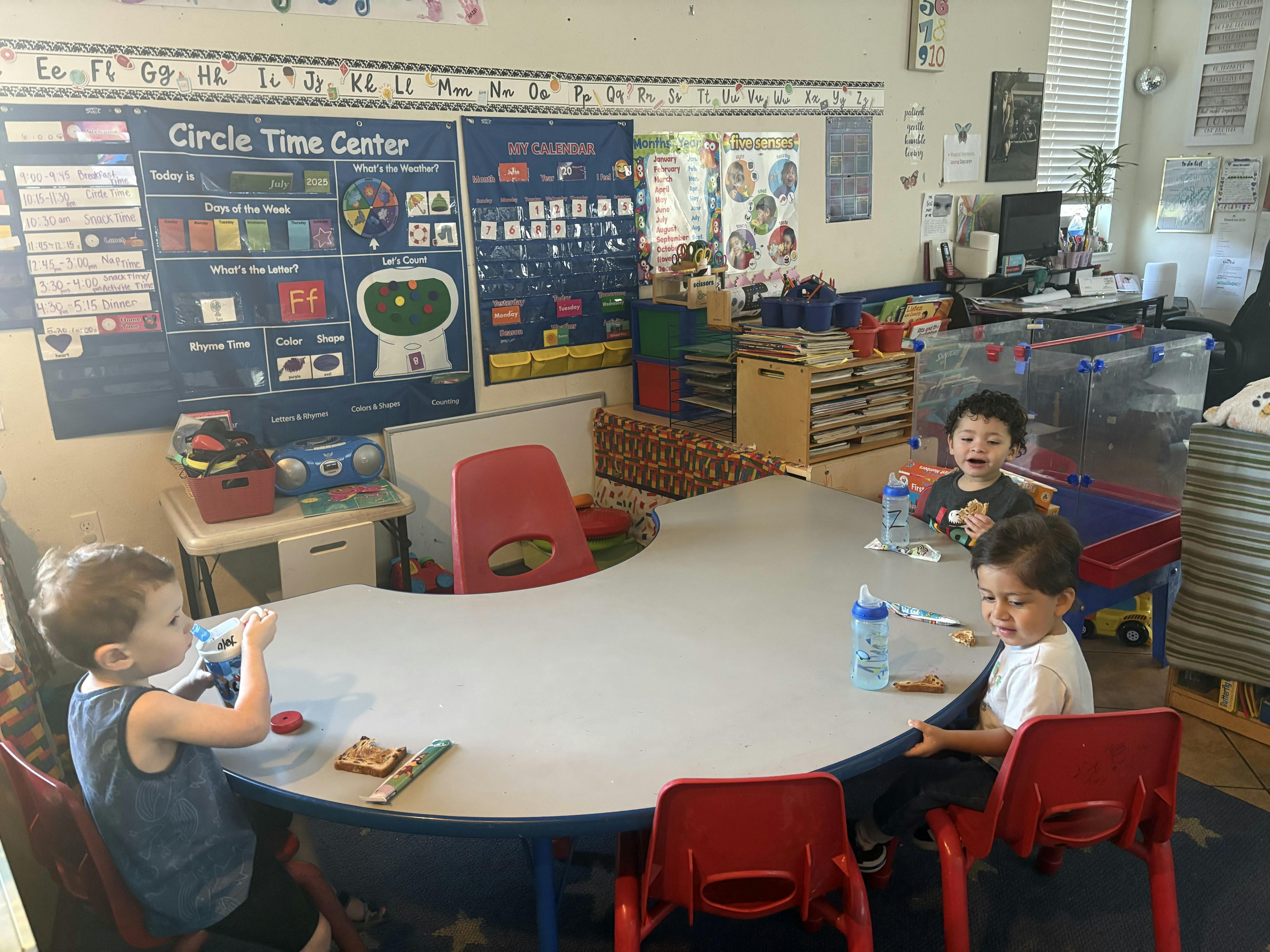 Three children sitting at a crescent-shaped table eating snacks in a classroom