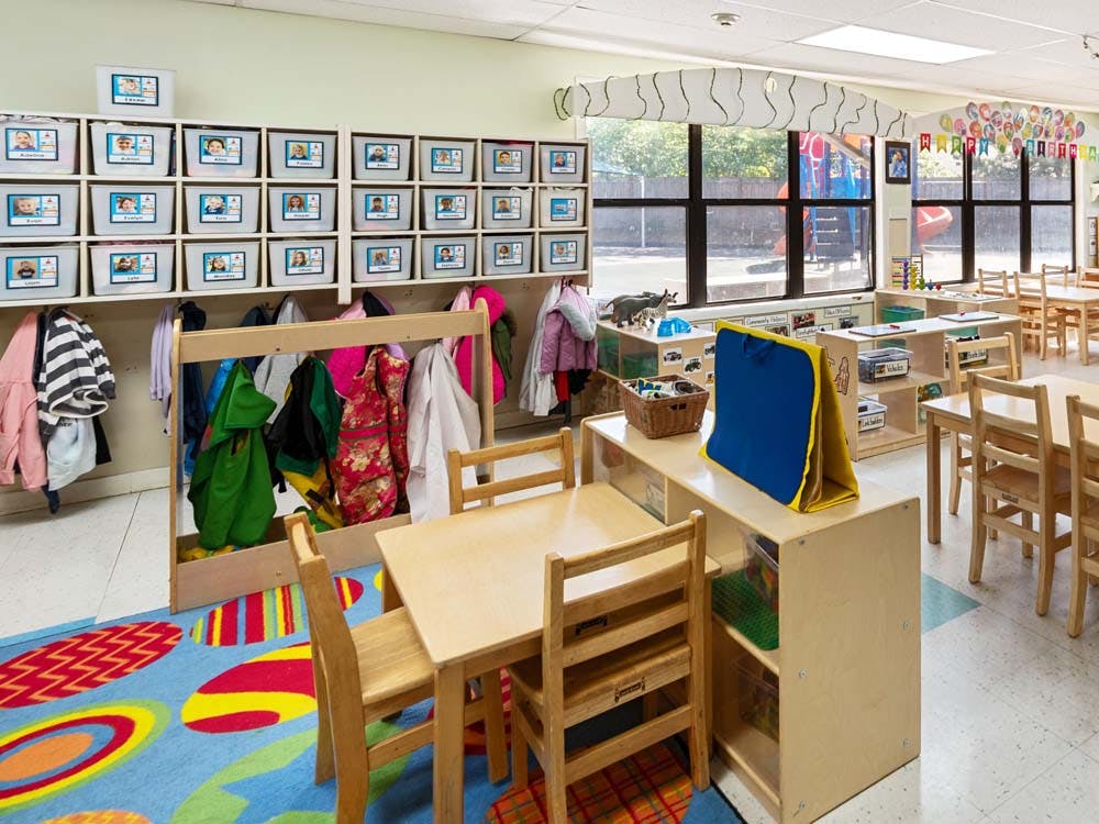 Empty classroom with small tables and chairs, coats hanging, and cubbies with student photos