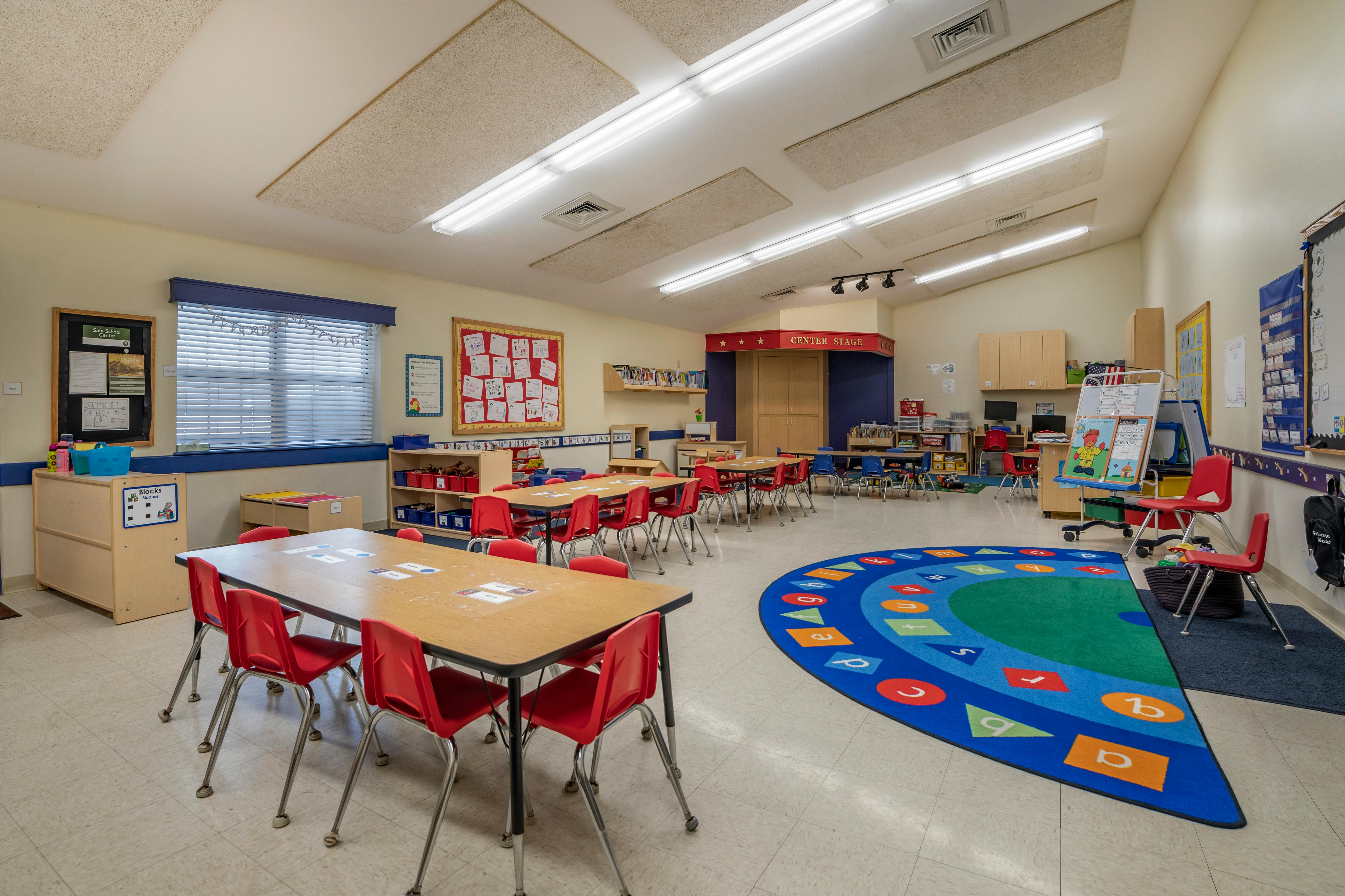 An empty classroom with small tables, red chairs, and a colorful alphabet rug