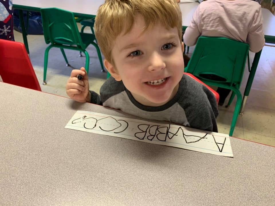Child holding a marker and looking at the camera, with letters on paper