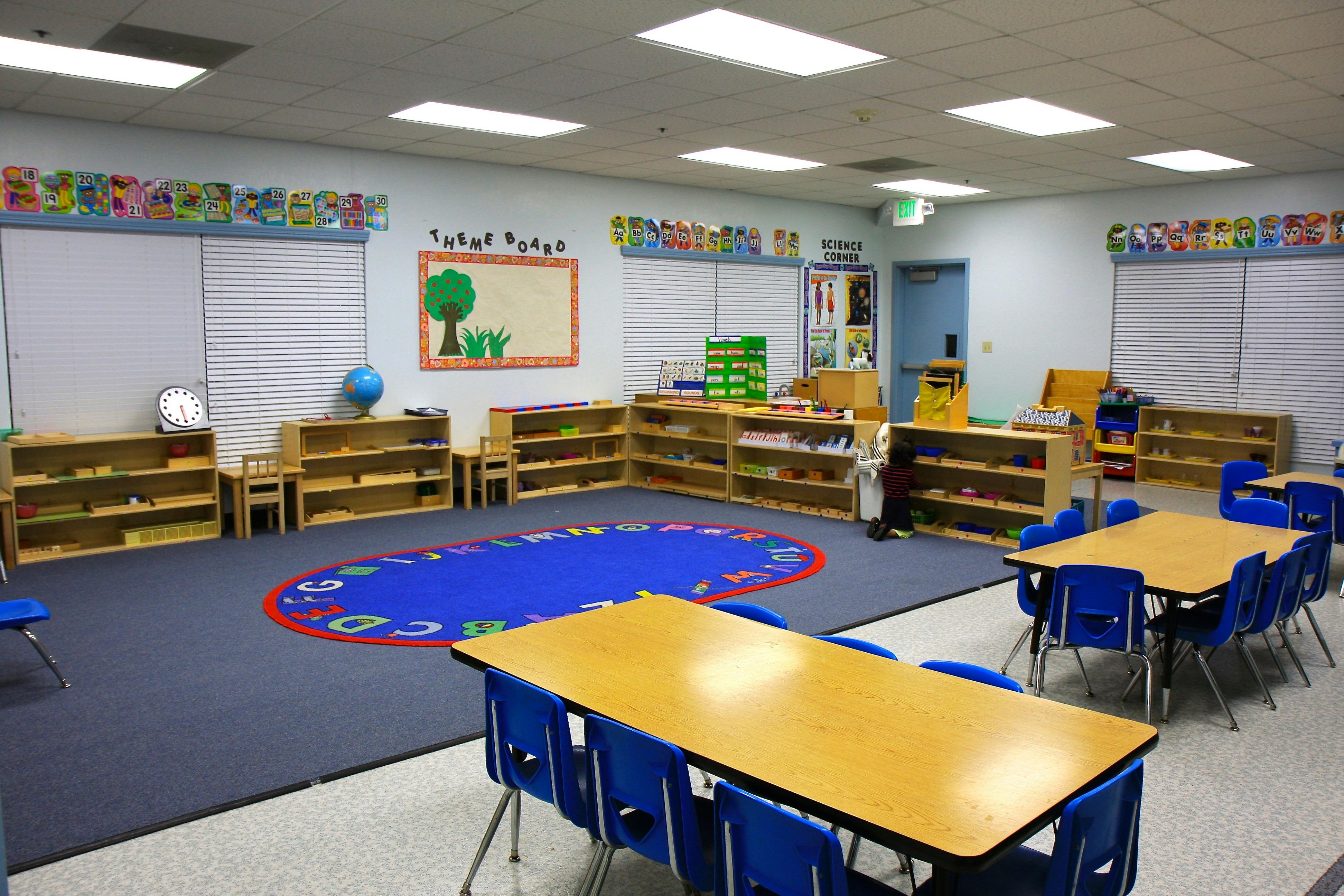 Classroom with small tables and chairs, shelves, and a blue oval rug