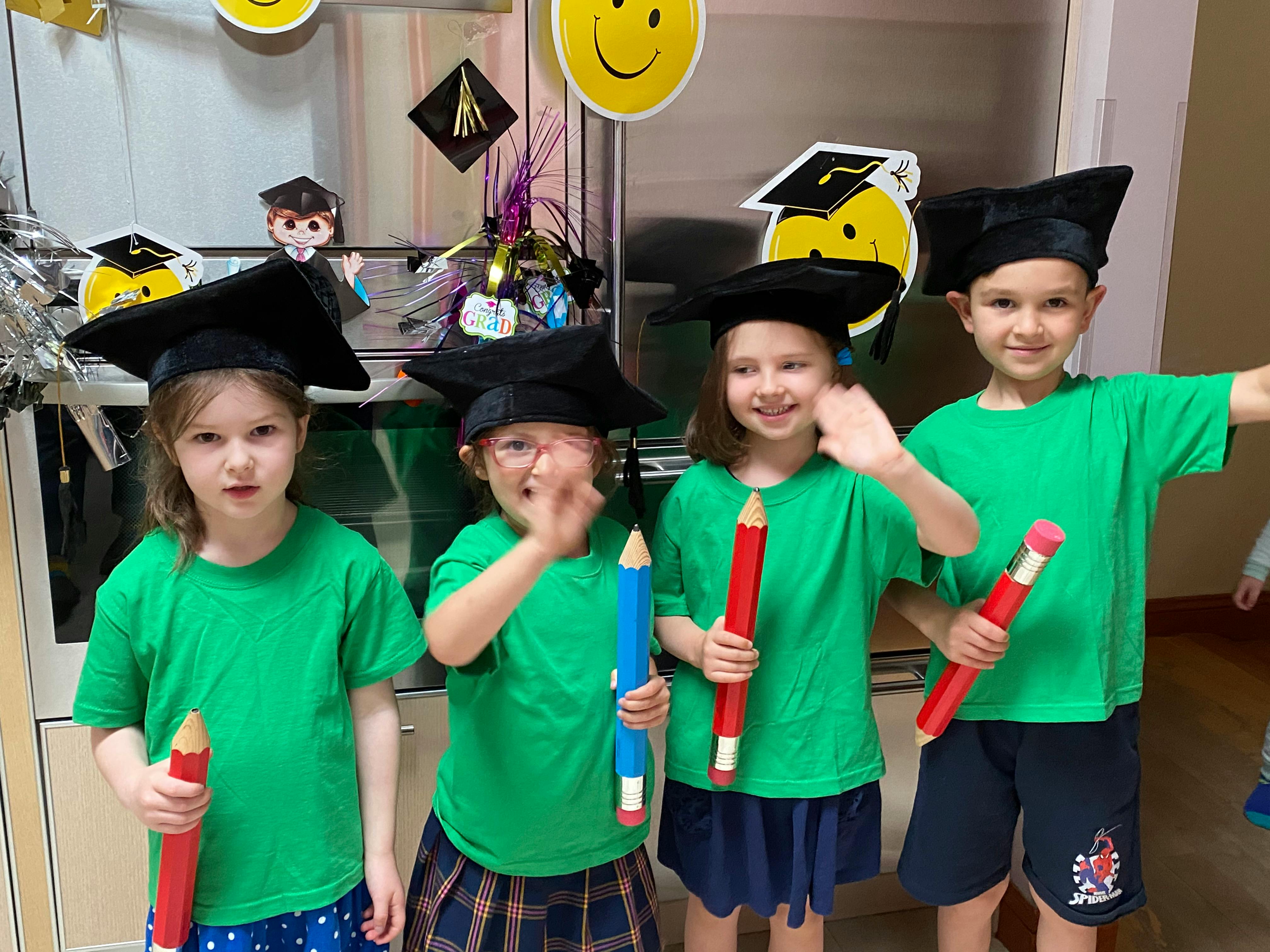 Four children in graduation caps and green shirts holding large pencils