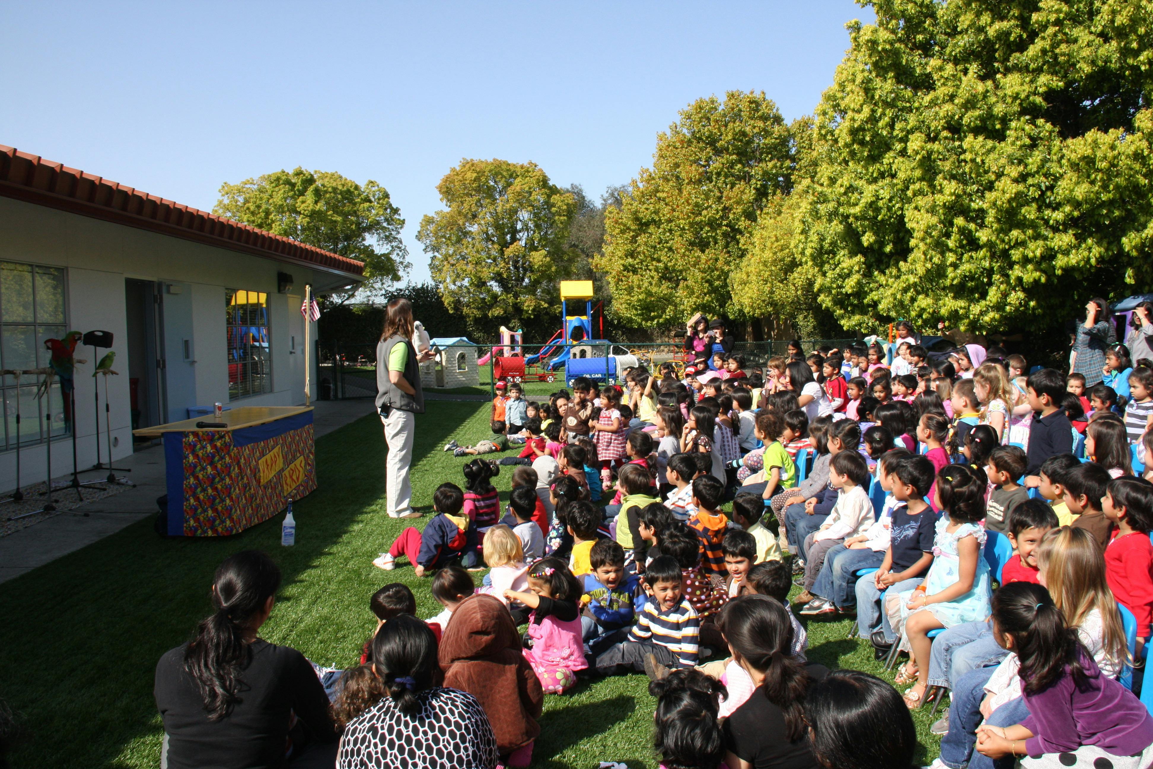 Person speaking to a large group of children and adults sitting on grass outdoors