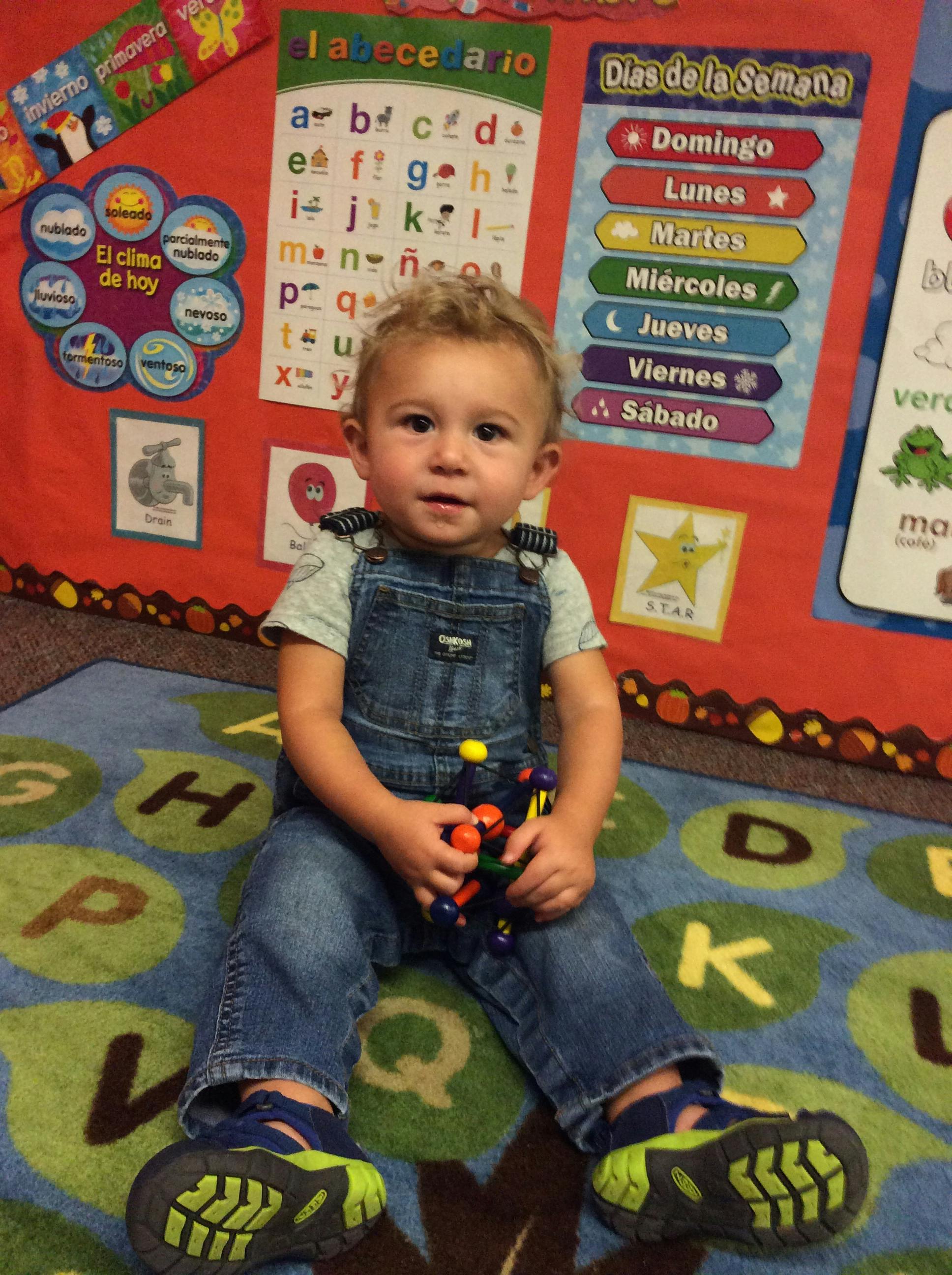 Child in denim overalls sitting on a lettered rug in a classroom