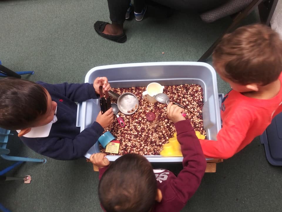 Three children playing with a sensory bin filled with beans and scoops