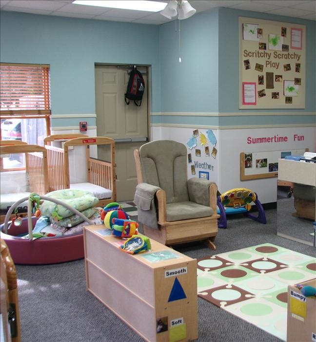 A childcare room with cribs, a rocking chair, play mat, and activity center