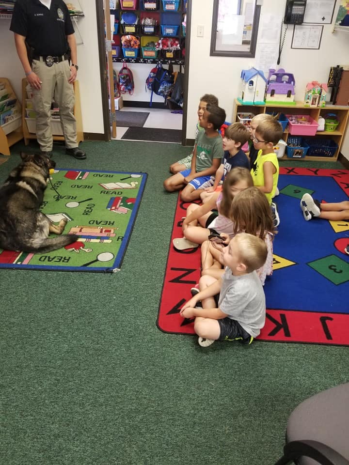 Person in uniform with a dog and children sitting on rugs in a classroom
